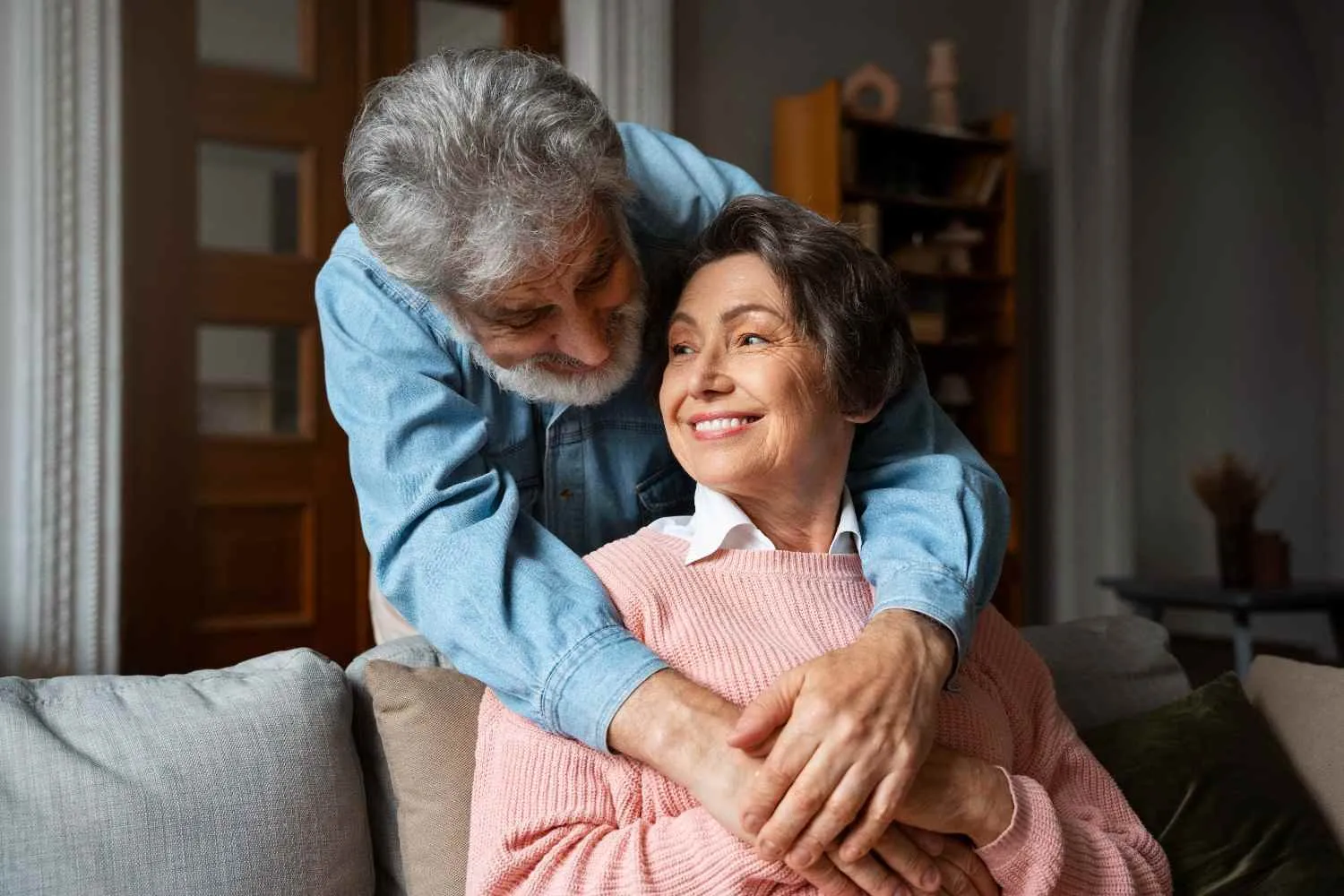 An older couple at home, one leaning over the sofa to hug the other